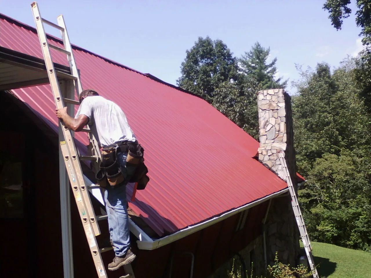 Roofer on a ladder beside a red metal roof and stone chimney — Soffit & Fascia Repair by Roofing Brothers in El Campo